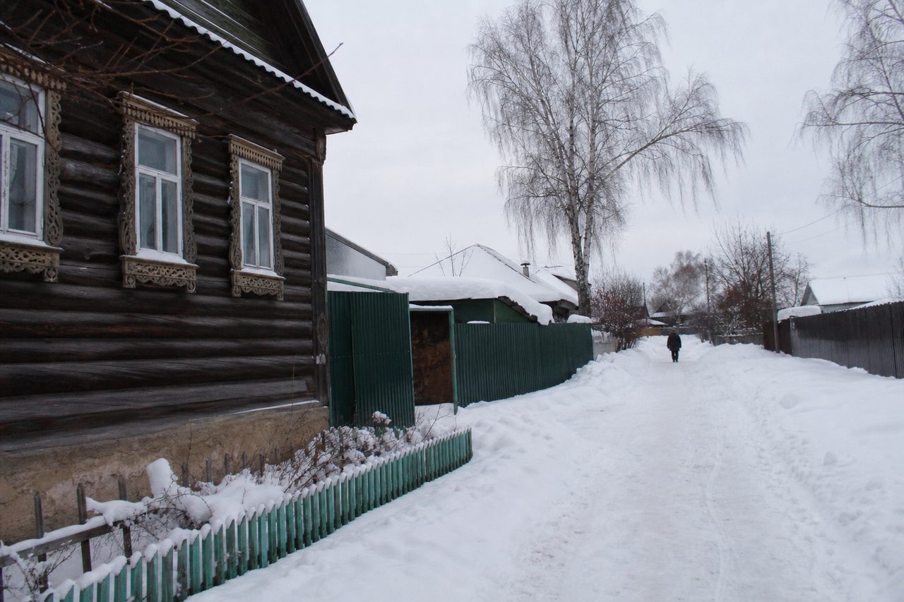 Заснеженная деревенская улица с деревянными избами, вдалеке по протоптанной тропинке уходит человек